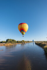 A vibrant multicolor hot air balloon flies low over the Rio Grande River, its reflection visible in the surface of the water. More balloons can be seen further upriver in the distance.