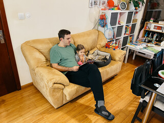 Dad reads a book to a little girl sitting on the couch next to a lying cat
