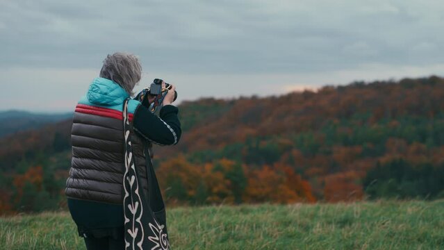 Cinematic shot of a woman photographer with grey hair taking pictures of an autumn landscape with her camera during a windy day surrounded by nature in slow motion. Shot with Sony FX3 in 4K