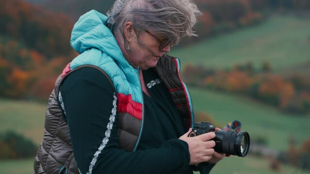Close up shot of a woman photographer looking at her camera, smiling and taking pictures, wind playing with her grey hair in an autumn landscape with colourful orange leaves. Sony FX3, slow motion, 4K