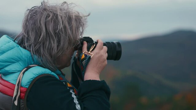 Cinematic close up shot of a woman photographer with grey hair taking pictures with her camera during a windy day in autumn nature in slow motion. Shot with Sony FX3 in 4K