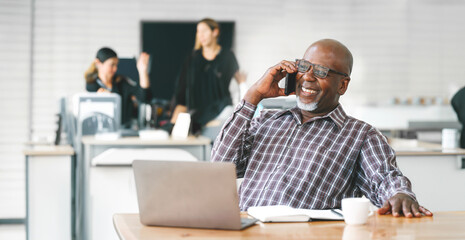 Portrait of happy African American small business owner talking on mobile phone. Millennial black male team leader smiling, employees working in modern office behind.