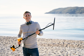 Smiling man with a metal detector at the ready stands by the sea