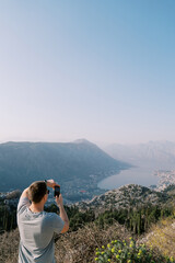 Man stands on a mountain and photographs a view of the Bay of Kotor with a smartphone. Montenegro. Back view