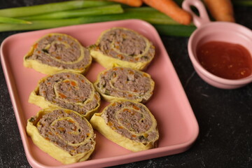 Rolade Daging Sapi. Beef roulade on a square pink ceramic plate. Made from ground beef, pepper, nutmeg, eggs, garlic, carrot, onion leaves. Served with tomato sauce.