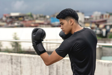 A young asian man makes an uppercut while practicing his boxing skills on the rooftop of a building. Shadowboxing training, wearing gloves, gritty urban scene. - obrazy, fototapety, plakaty