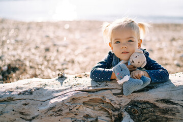 Little girl sits on the beach leaning on a snag in an embrace with soft toys
