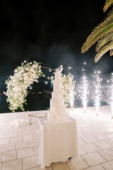 Multi-tiered wedding cake stands on a table near a round wedding arch in the light of fireworks