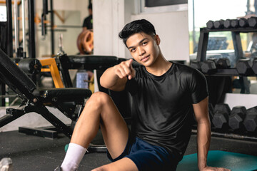 Fototapeta na wymiar A young handsome and confident asian man pointing at the camera while sitting on the mat at the a popular open air gym. Choosing you as a training partner.