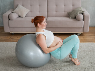 Happy pregnant woman lies with her back on a fitball and keeps her hands on her tummy at home.