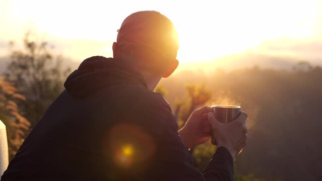 Hiker man sitting on chair, holding coffee mug with steam at dawn. Back view of young traveler that woke up early morning to meet dawn and bathe in sunshine. Concep travel on camping van to meet dawn.