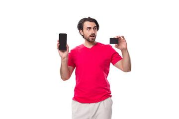 young surprised male promoter dressed in a red basic t-shirt holds a plastic card and a mobile phone