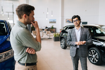 Rear view of stylish male car dealer in business suit having conversation with pensive young man client at showroom. Doubtful customer guy listening manager while buying car at dealership center - Powered by Adobe