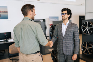 Naklejka na ściany i meble Rear view of happy customer male thanking stylish dealer male wearing suit and eyeglasses for helping to choose car to buy in dealership center. Concept of choosing and buying new car at showroom