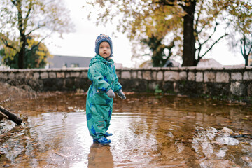 Little girl in rubber boots stands in a puddle in an autumn park. Side view