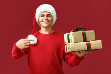 Young man in Santa hat pointing at gift boxes on red background
