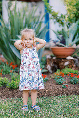 Little girl stands thoughtfully in the garden by the flower bed, touching her face with her hands