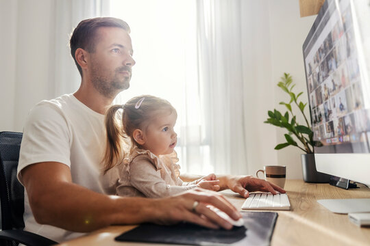 A stay at home dad is working on a computer at his home office while babysitting his daughter.