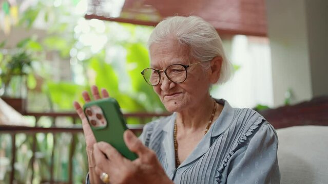 Happy Asian grandmother in eyeglasses video call with family through smartphone. Elderly woman in eyeglasses waving hand video call distant conversation chatting online at home. Technology concept
