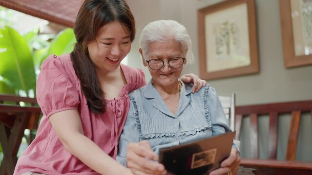 Asian grandmother and young granddaugther looking through a family photo album together while leisure time at home. Family moments and memory of life. Family Relationship concept
