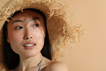 Beautiful young woman in straw hat with sun protection cream on her face against beige background,...