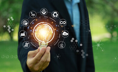 Environmental protection, renewable, sustainable energy sources.businessman holding light bulb against nature on a green background with energy source icons, sustainable development, ESG, Ecology - obrazy, fototapety, plakaty