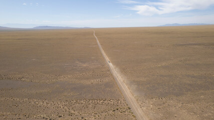 A white SUV is driving fast on a dusty road in the steppe. Clouds of dust fly from under the wheels and from the roof of the car. The jeep easily passes a dirt road. Blue sky and white clouds.