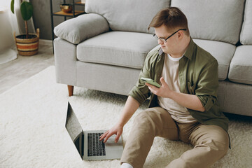 Young man with down syndrome wears casual clothes use laptop pc computer mobile cell phone sit grey sofa couch stay at home flat rest spend free time in living room. Genetic disease world day concept. - obrazy, fototapety, plakaty