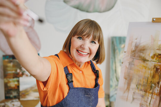 Close up elderly artist woman 50 years old wearing casual clothes stand near easel with painting stretch hand with brush to camera spend free spare time in living room indoor. Leisure hobby concept.