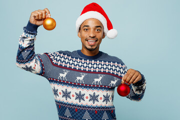 Young smiling man wear knitted sweater Santa hat posing hold in hand two toys for Christmas tree look camera isolated on plain pastel blue background. Happy New Year 2024 celebration holiday concept.