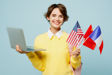 Young happy woman IT student wear casual clothes yellow sweater backpack bag hold many flags use work on laptop pc computer isolated on plain blue background. High school university college concept.