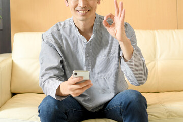 スマートフォンを持ちながらOKサインをする私服姿の若いアジア人男性
