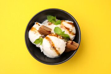 Tasty ice cream with caramel sauce, mint, candies and nuts in bowl on yellow background, top view
