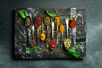 A set of colored spices in metal spoons. On a black slate background. Top view.