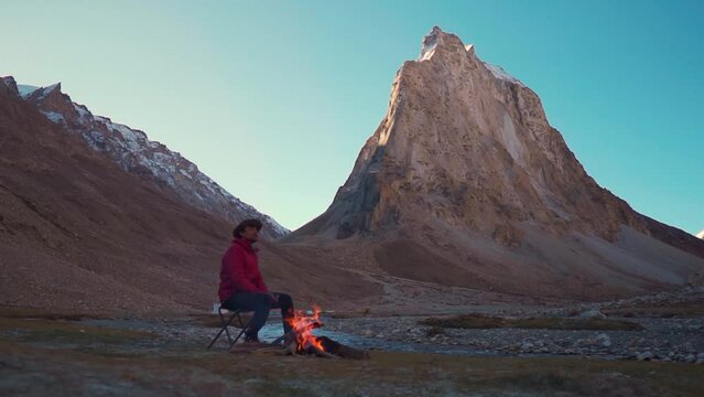 Happy Indian male tourist sitting besides the bonfire in front of the Holy Gonbo Rangjon mountain in Zanskar Valley at Ladakh, India. Tourist warming himself with fire in cold Himalayas in India.