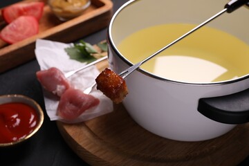 Fondue pot, forks with meat pieces and other products on black textured table, closeup