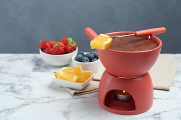 Fondue pot with melted chocolate, fresh orange, different berries and fork on white marble table. Space for text