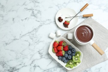 Fondue pot with melted chocolate, sweet marshmallows, fresh kiwi, different berries and forks on white marble table, flat lay. Space for text
