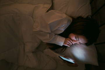 Portrait of Asian teen girl lying in bed at night and using smartphone. Young woman lying in bed using smartphone for social media and watching series at late night. Insomnia, sleep disorder concept.