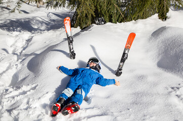 love winter, cheerful preschooler boy in blue overalls lies on snow, skis next to him. Seasonal joys, happy childhood. Frosty weather. Children's skiing lesson at ski school, break. sports training