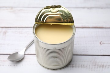 Tasty condensed milk in tin can and spoon on white wooden table, closeup