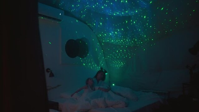 Woman with children looks at bright starry sky glowing on ceiling sitting on bed in modern room. Dim light source making lighting in premise soft and soothing slow motion