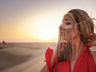 Young arabian Woman in red silk dress with gold veil accessories hide face. Oriental beauty fashion model. sands dunes of UAE desert at sunset. Dubai Desert Conservation Reserve, United Arab Emirates