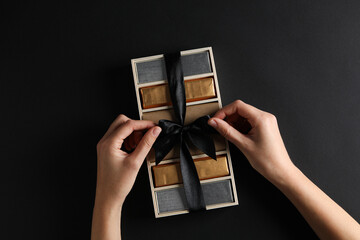 Chocolates in box with ribbon and hands on black background, top view