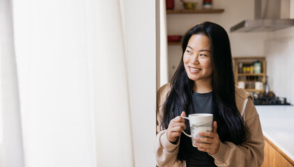 Young beautiful woman with a mug drinking coffee next to a window in a kitchen. Smiling woman...
