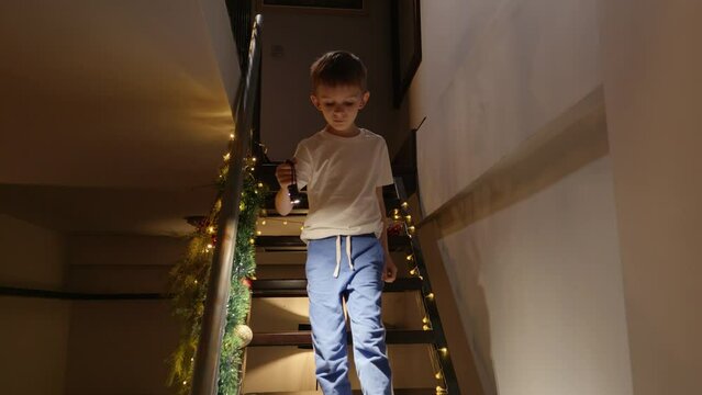 Excited little boy waiting for Santa Claus on Christmas eve walking down the stairs at house with flashlight. Children celebrating winter holidays