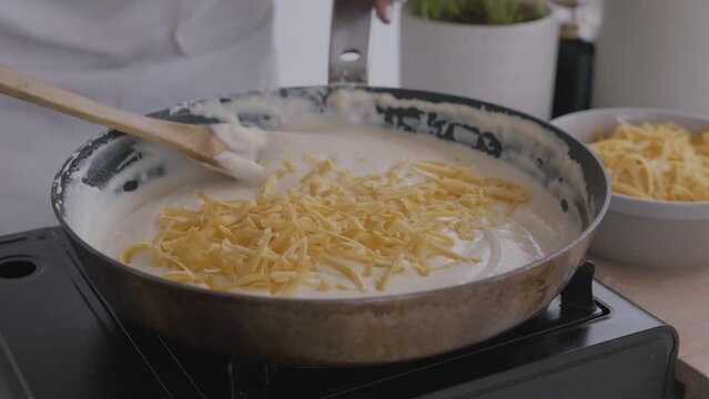 A woman cheese and b&eacute;chamel in a frying pan with wooden spoon. Preparation of cheese sauce for croutons or macaroni and cheese. Side view of kitchen table. Cooking in the kitchen. Delicious cooking.