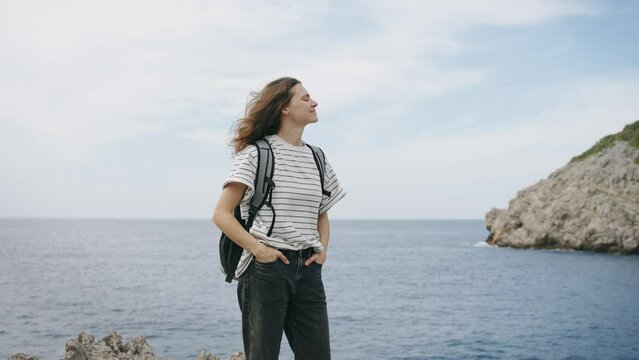 A young tourist with a backpack enjoys the view while standing on top of a rocky shore overlooking the sea