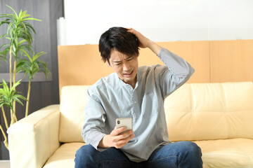 頭を抱えながらスマートフォンを見るアジア人男性
