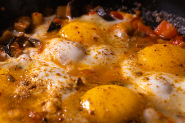 Close-up of fried chicken eggs, tomatoes and eggplant with seasoning in a frying pan. Cooking process, selective focus. Homemade food for breakfast